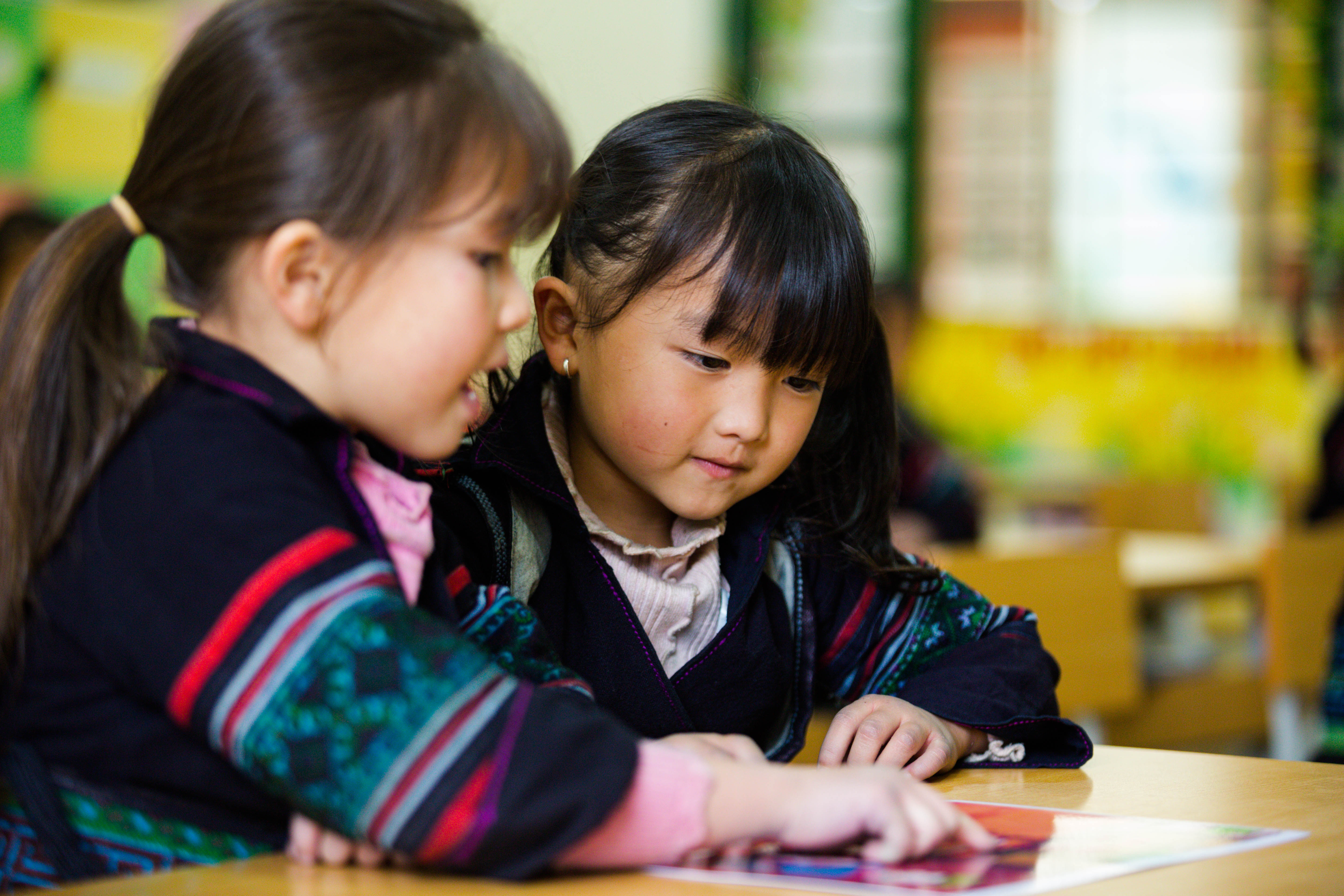 Two six-year-old girls sit at a desk in a classroom reading and pointing to a pamphlet.