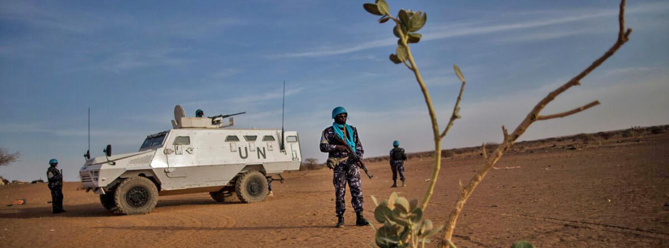 Un miembro del personal de mantenimiento de la paz de la ONU haciendo guardia con 2 miembros del personal de mantenimiento de la paz y un vehculo de la ONU al fondo.